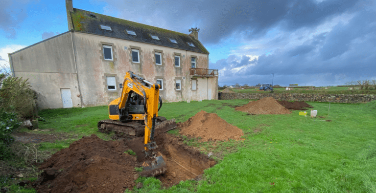 Mini-pelle JCB réalisant des sondages de sol et tests d'infiltration pour une étude d'assainissement non collectif devant une maison en pierre bretonne